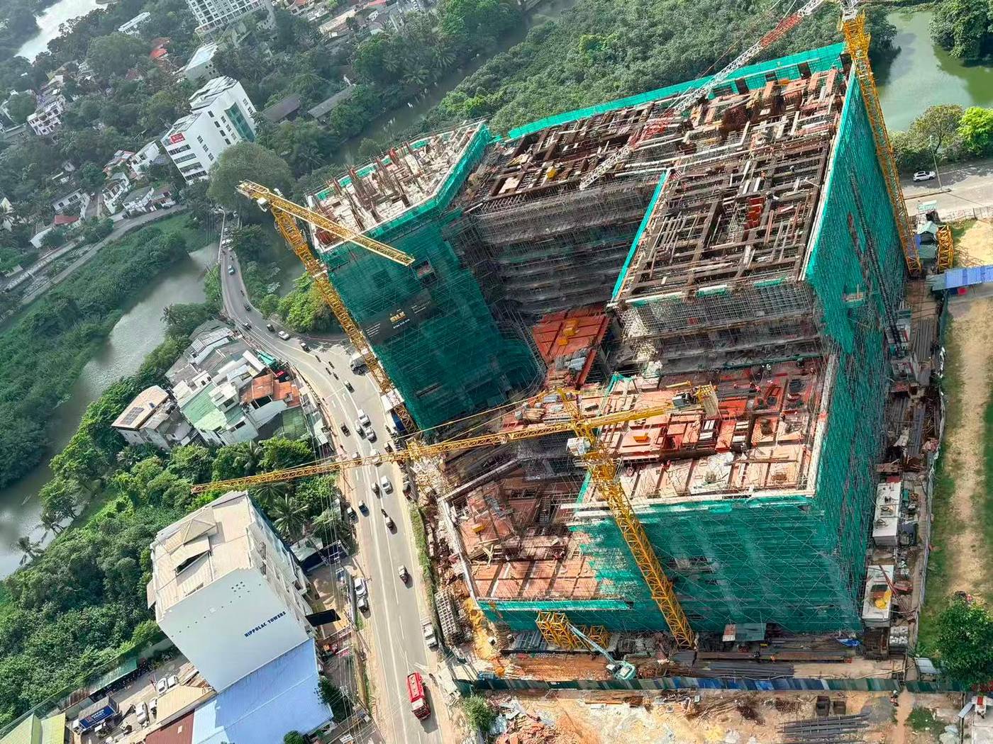 Aerial view of the SILK development under construction in Rajagiriya, green safety netting across the frame, two tower cranes, with branded hoarding visible on the structure and the Diyawanna lake beyond.